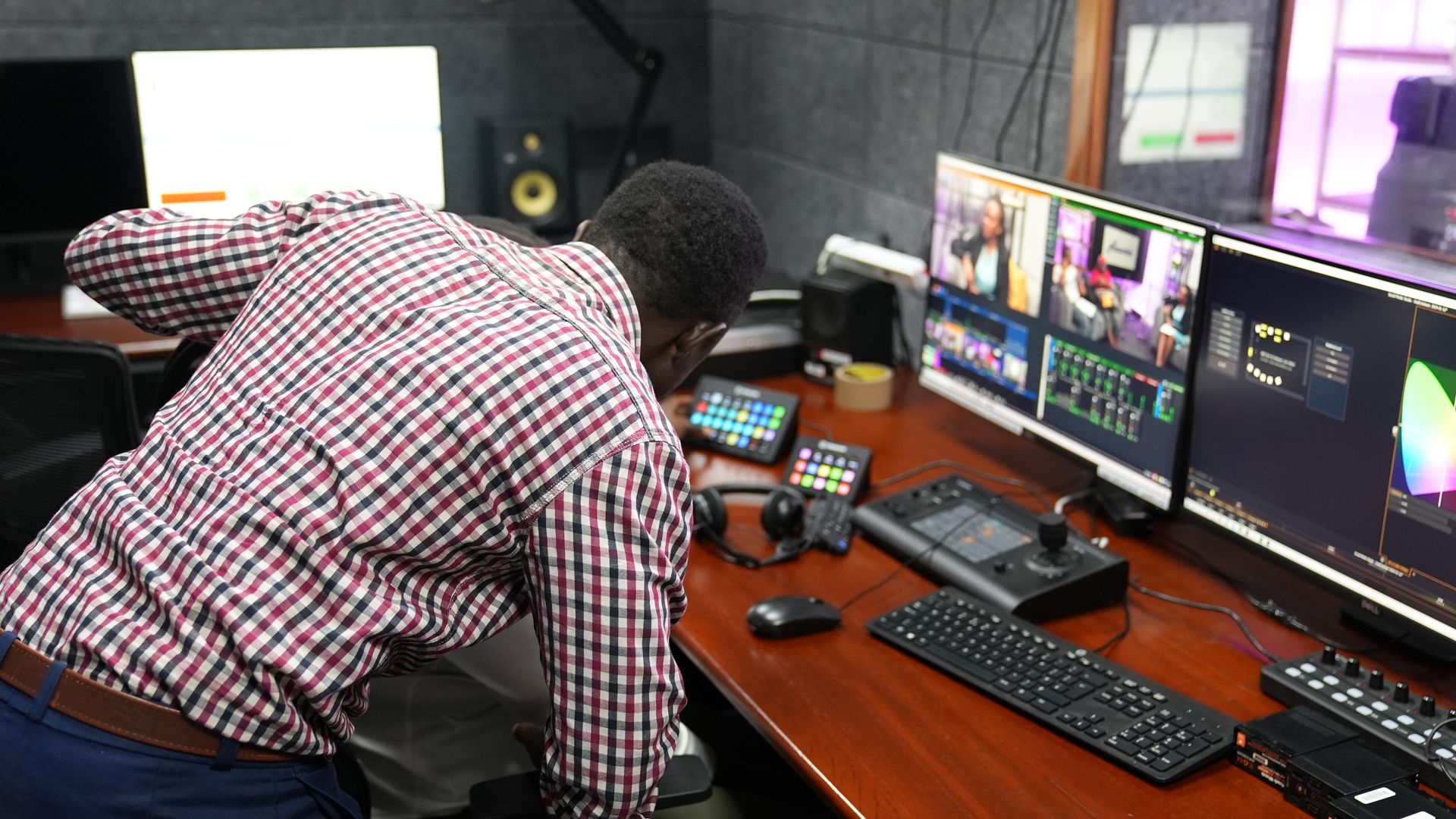 A man in a checked shirt is standing over a chair where someone is sitting out of shot. Technical equipment is spread on top of a desk next to them.