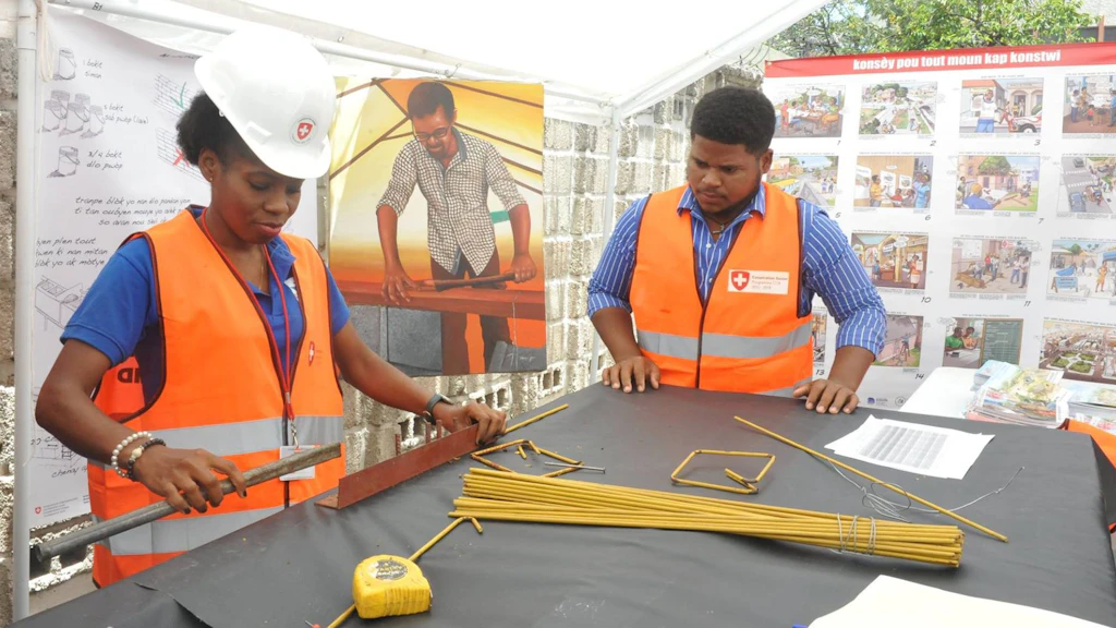 A trainer observes a trainee working with metal bars and construction plans. They are wearing reflective safety vests and hard hats.