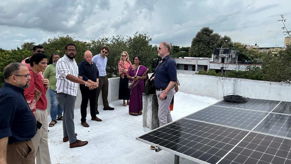A group of about ten people stand on a flat rooftop fitted with solar panels under a cloudy sky, engaged in conversation.