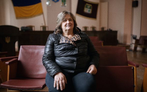 Liudmyla is sitting in her living room, with a Ukrainian flag hanging in the background.