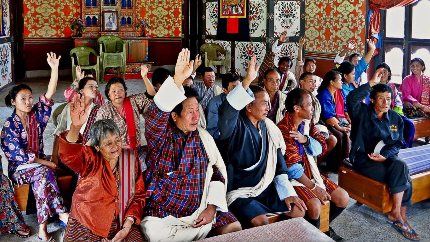 Citizens of all ages, sitting on benches in a rural community in Bangladesh, engage in the Local Governance planning process by voting with raised hands.