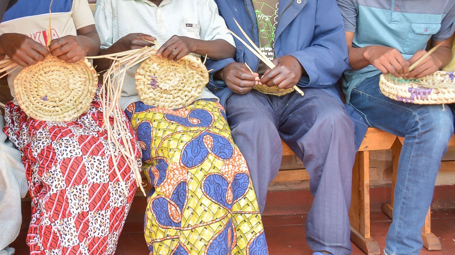 Five men and women are sitting next to each other, looking down on the baskets they are weaving. A woman is leaning towards them.
