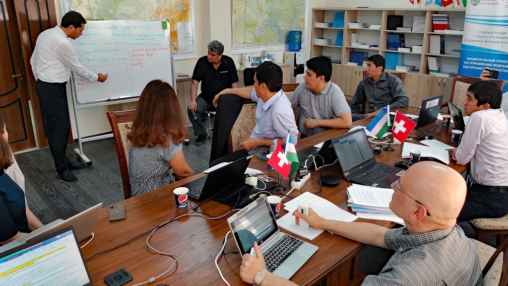 People are sitting at a table. A man is presenting at a flipchart, maps are hanging on the wall, and laptops and flags of Switzerland and Uzbekistan are on the table.