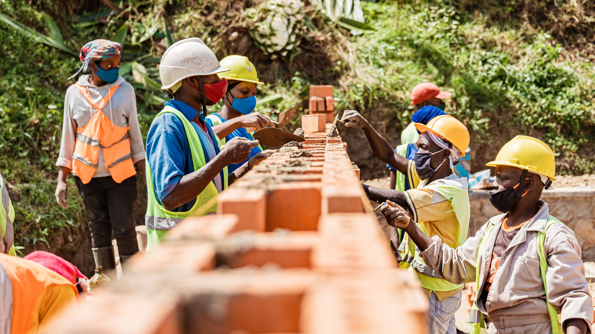 Various people in workers' clothing, wearing high-vis jackets and hard hats, are building a brick wall.