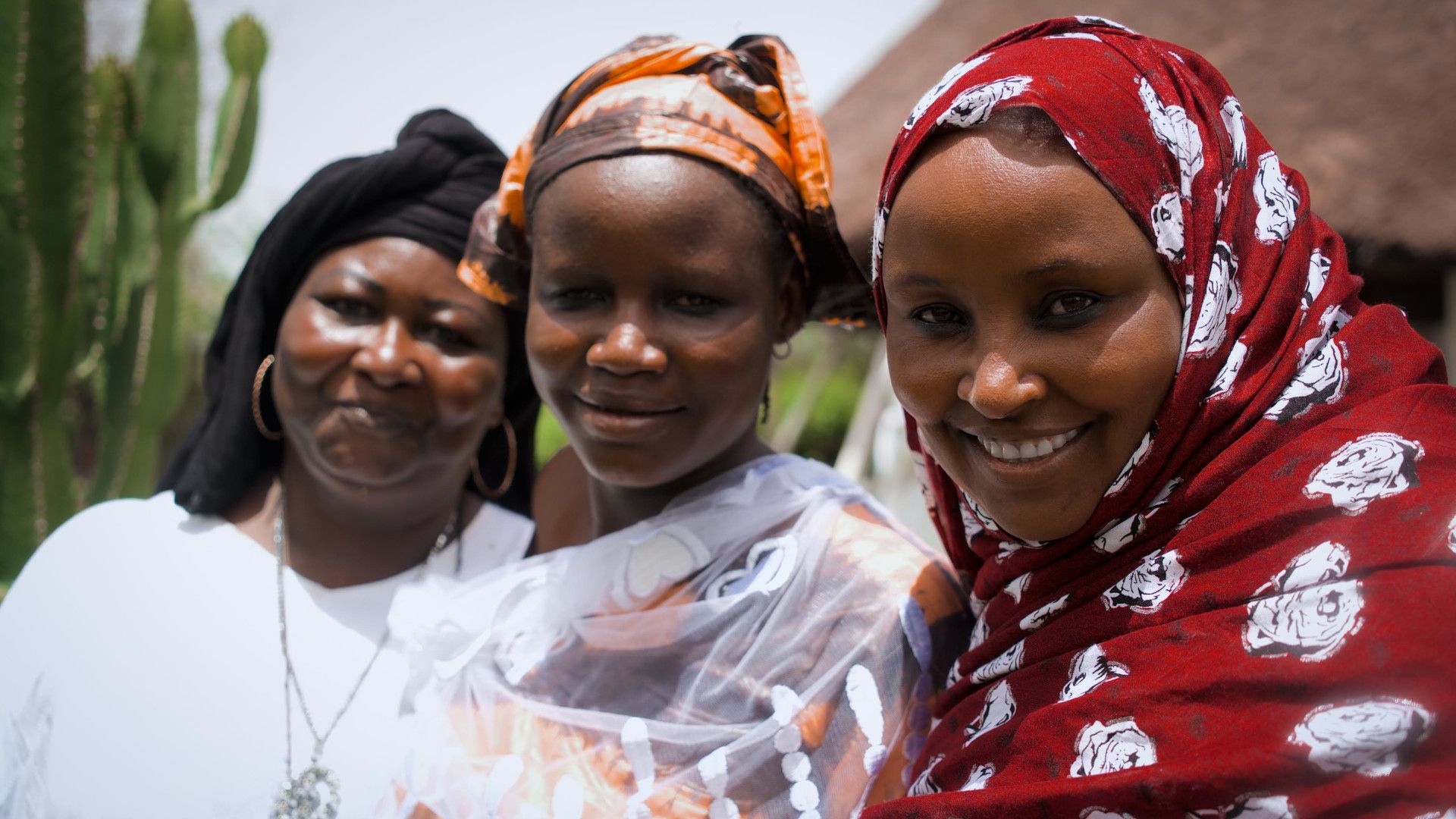 Three young women wearing white and coloured clothing standing side by side, smiling.  