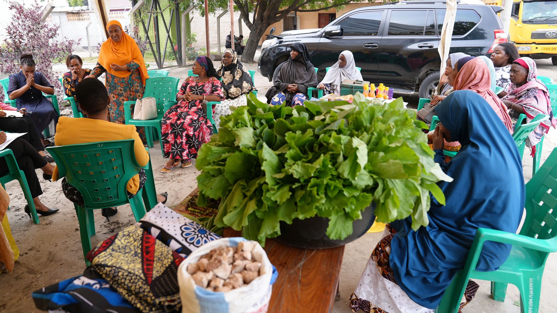 A group of women are sitting in light green chairs listening to a woman who is standing. In the foreground garments, lettuce and a bag of stones are laid out on a table.