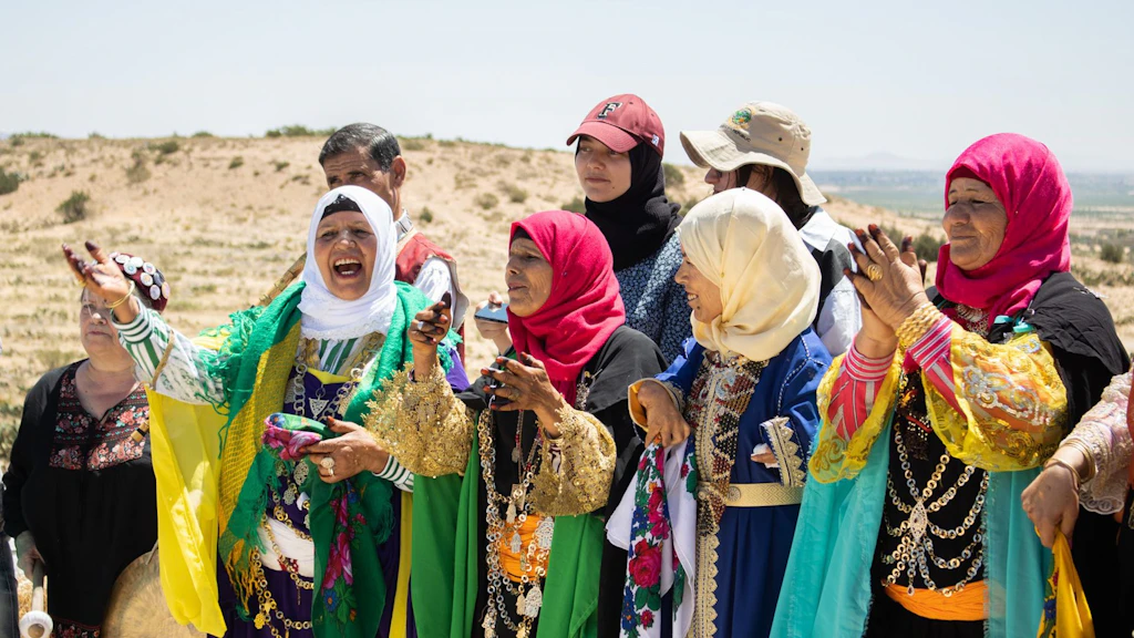 Women in colorful traditional clothing sing and clap in a desert landscape. Some wear bright headscarves and jewelry.
