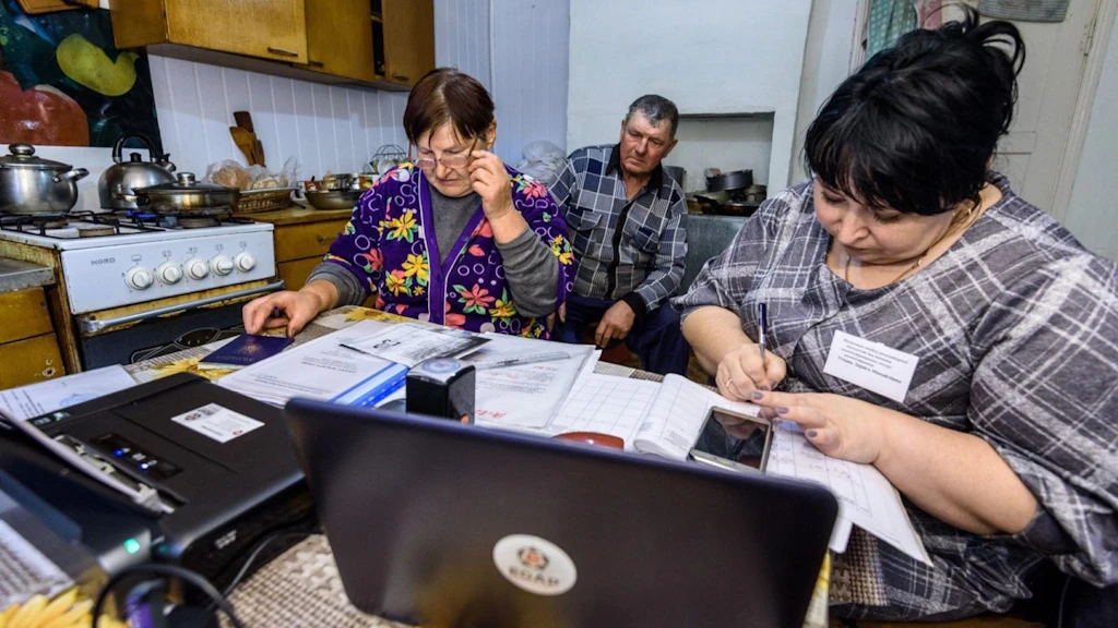 Three people sitting in a kitchen working on documents on a laptop.
