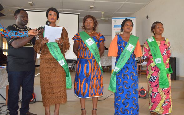 Four women in colourful dress. The first is holding some sheets of paper and a man is handing her a microphone.