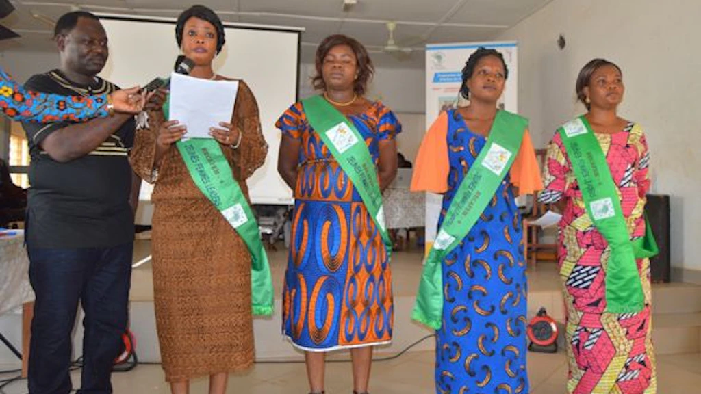 Four women in colourful dress. The first is holding some sheets of paper and a man is handing her a microphone.