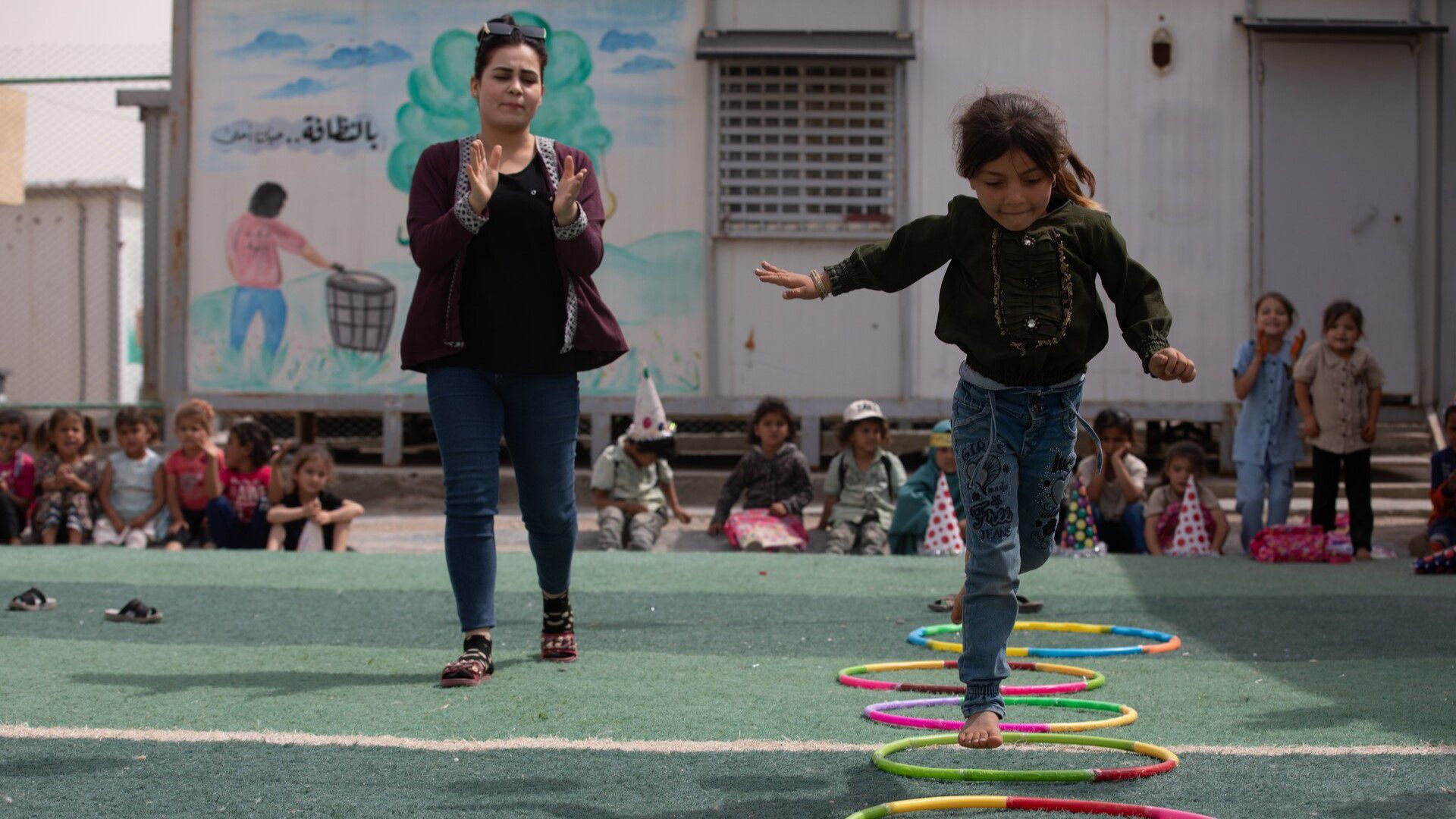 A young girl plays in a school playground with a teacher who applauds her from the side.