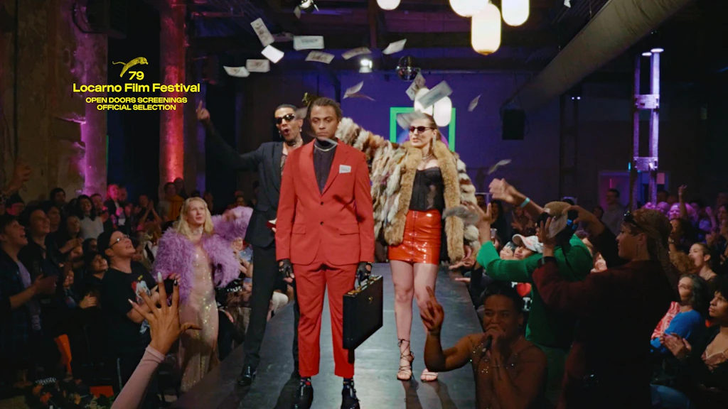 A young man in a red suit is standing on a podium, surrounded by a lively audience and banknotes flying around him. 