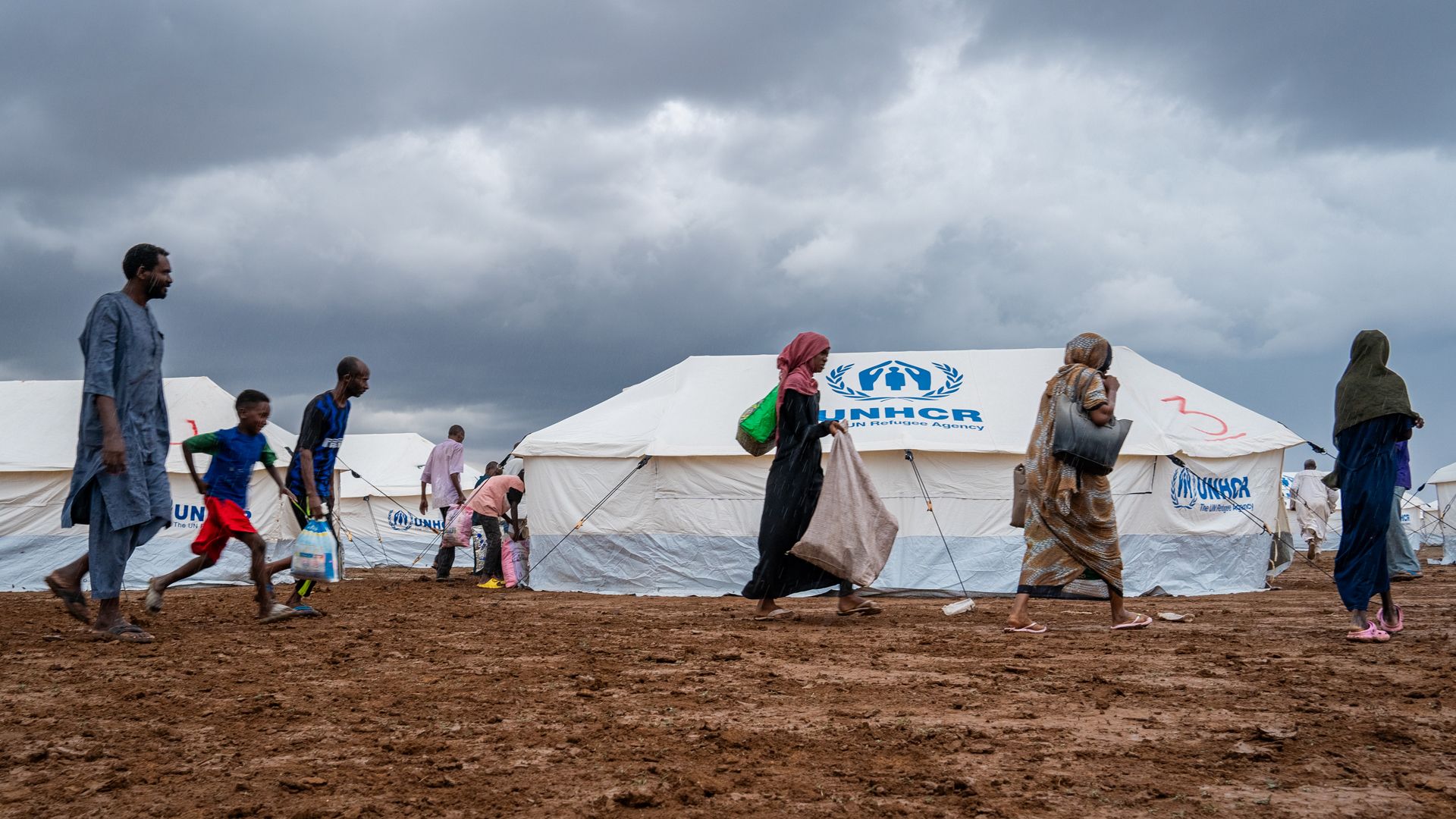Several people walk past a white tent. The logo of the United Nations Refugee Agency, printed in blue, can be seen on the tent.