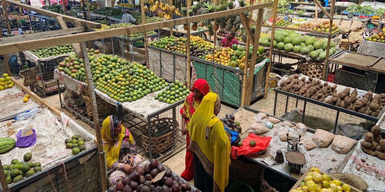 Two women in red and yellow veils stand in front of their fruit stall. Shelves full of fruit and vegetables surround them.