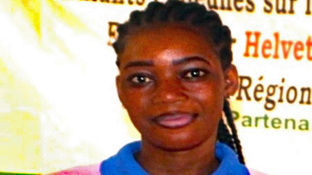 A young woman in a pink T-shirt with a blue collar, with glasses hanging from her T-shirt, looks straight ahead.