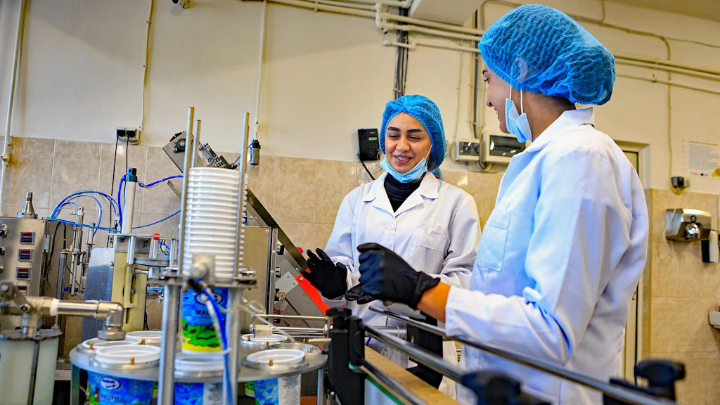 Two young women in Armenia standing by a filling machine for dairy products and gaining hands-on experience of food safety and quality control.