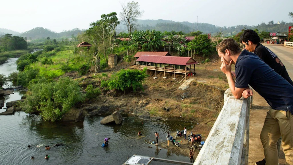 Two people are leaning on the side of a bridge, with people below them working to set up a sensor in the river.
