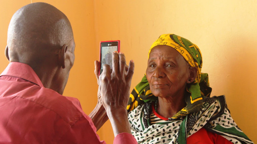 A man takes a portrait photo with his mobile phone of a woman, who is standing in front of an orange wall.