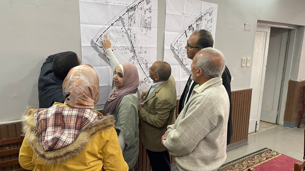 A group of women and men are looking at an urban development plan displayed on the wall. One woman in the group is pointing to a spot on the plan.