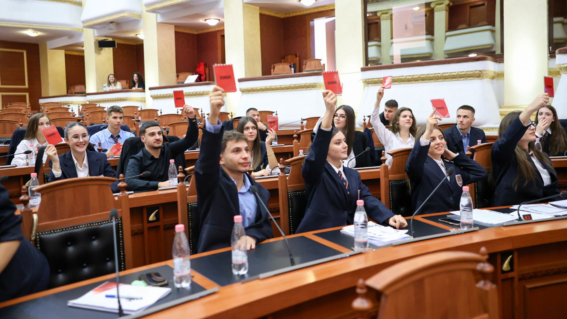Young participants simulate a vote by raising red notepads in a parliamentary chamber. 