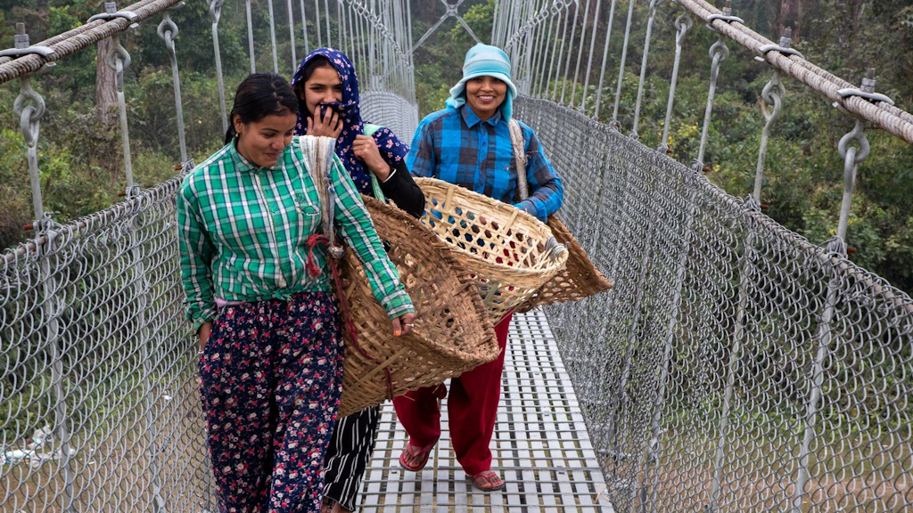 Three Nepalese women cross a suspension bridge. They are carrying large, empty wicker baskets.
