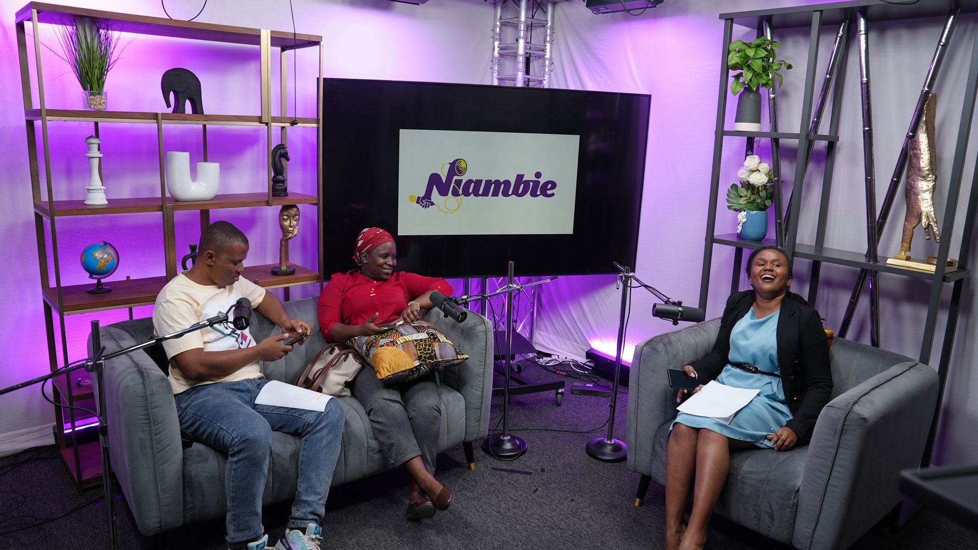 Two people are sitting on a sofa, another person is sitting on a chair laughing. They are in a recording studio with “Niambie” written on a large TV in the background.