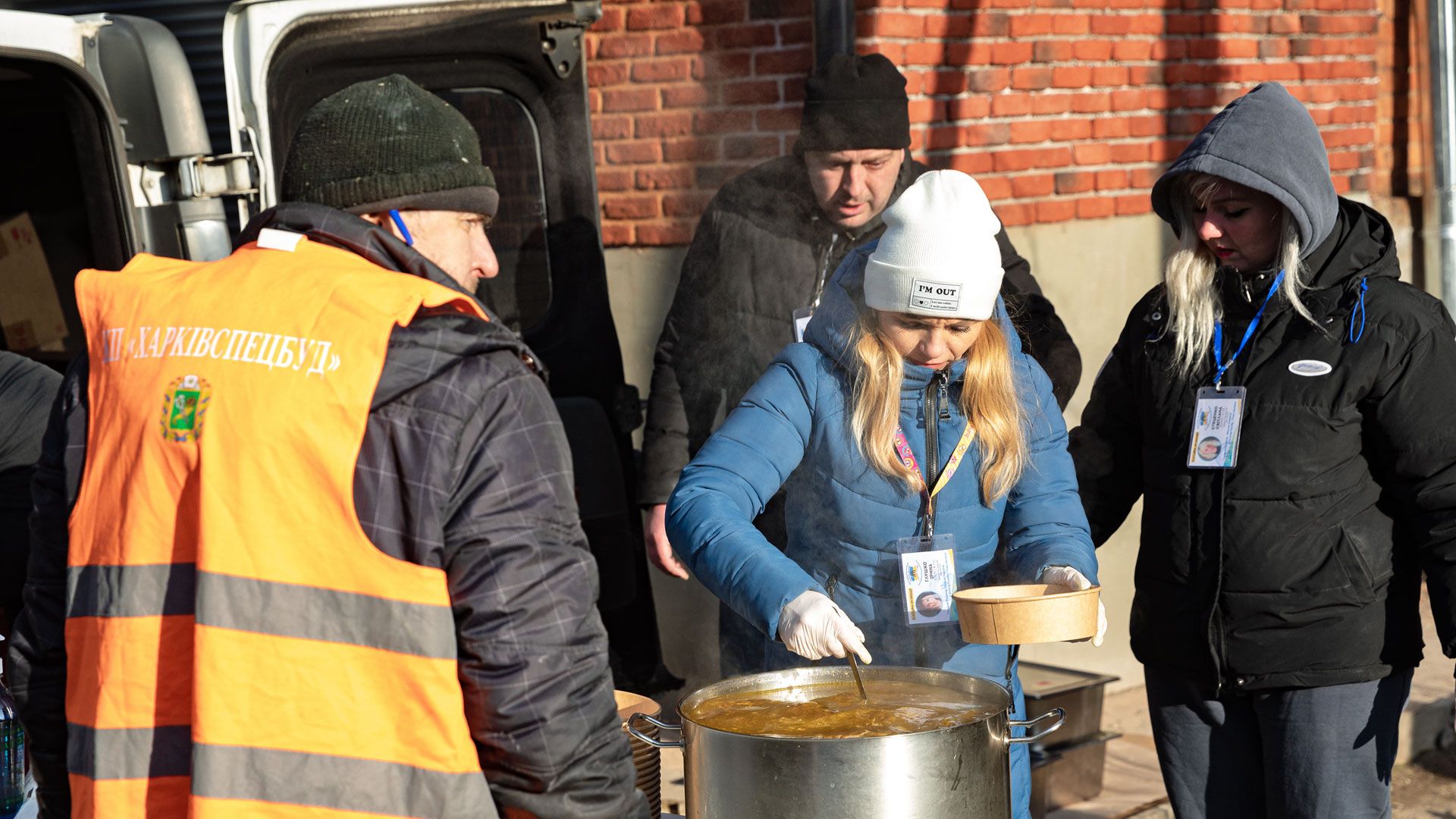 Volunteers handing out hot soup outside on a frosty day. Steam rising from the large pot by a brick wall.