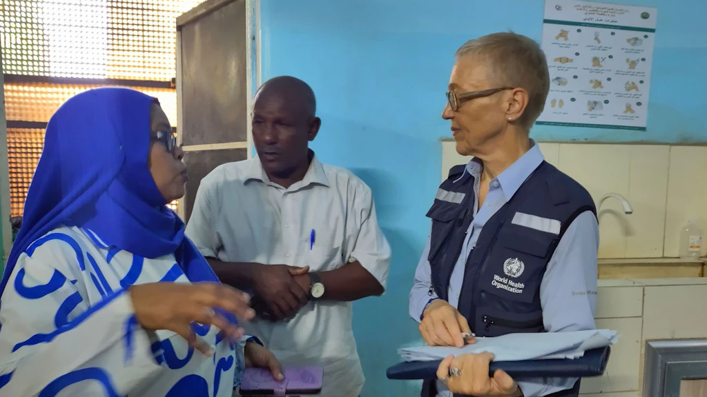 Une femme portant un foulard bleu s’entretient avec une experte de l’OMS, qui tient des documents, Un homme se tient à leur côté.
