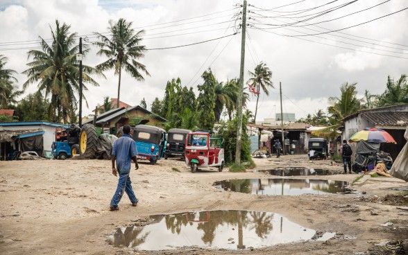 Un homme vêtu d’un bleu de travail marche dans une rue jonchée de nids-de-poule remplis d’eau. Des véhicules isolés longent le bord de la route, qui est par ailleurs vide.