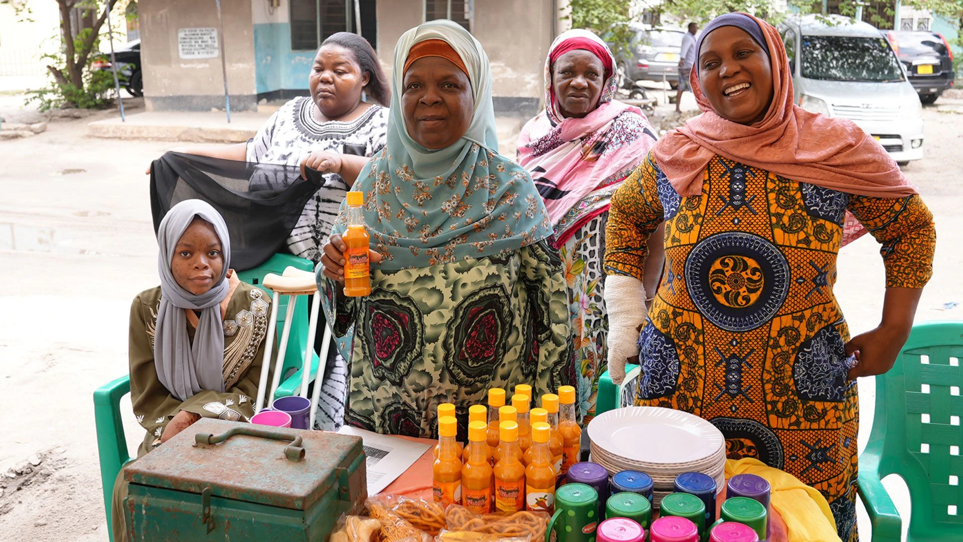 Cinq femmes, quatre debout et une assise, se tiennent derrière une table. Des mugs, des assiettes, des noix emballées dans des sacs plastique et des bouteilles de sauce au piment sont posées dessus. 
