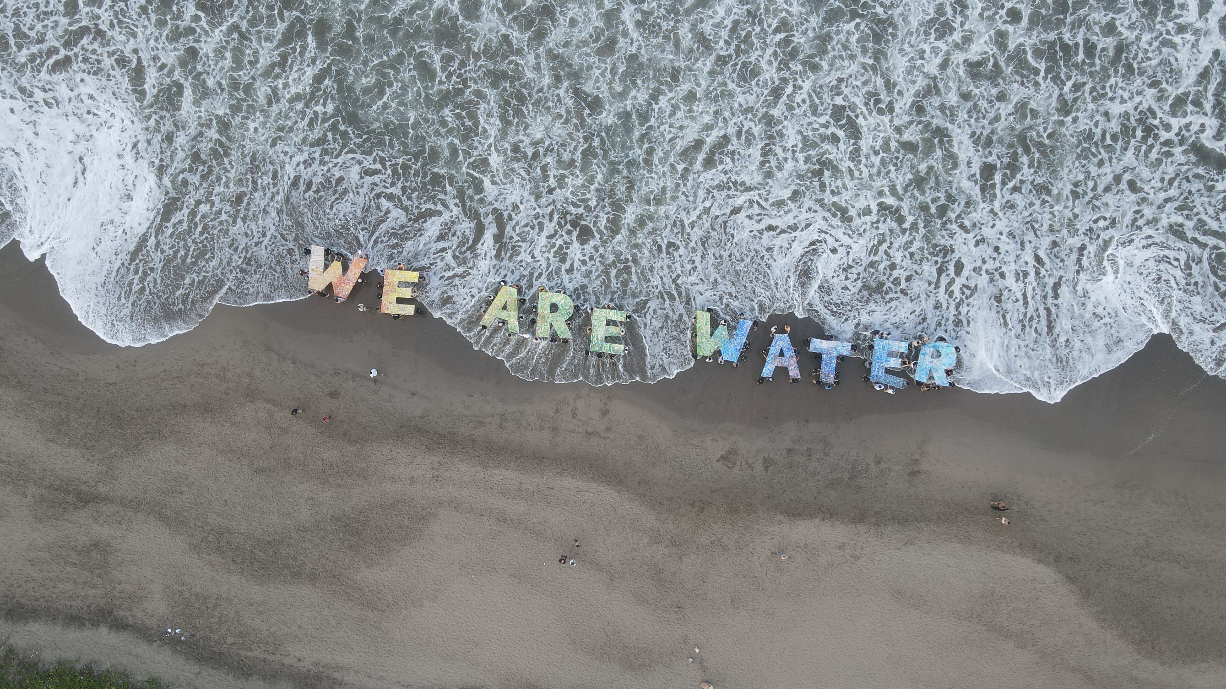 Depuis un point de vue élevé, on aperçoit une plage de sable baignée par les vagues de l'océan. Sur le sable, un message coloré et imposant, 'We Are Water', apparait. Il est très coloré et captivant.