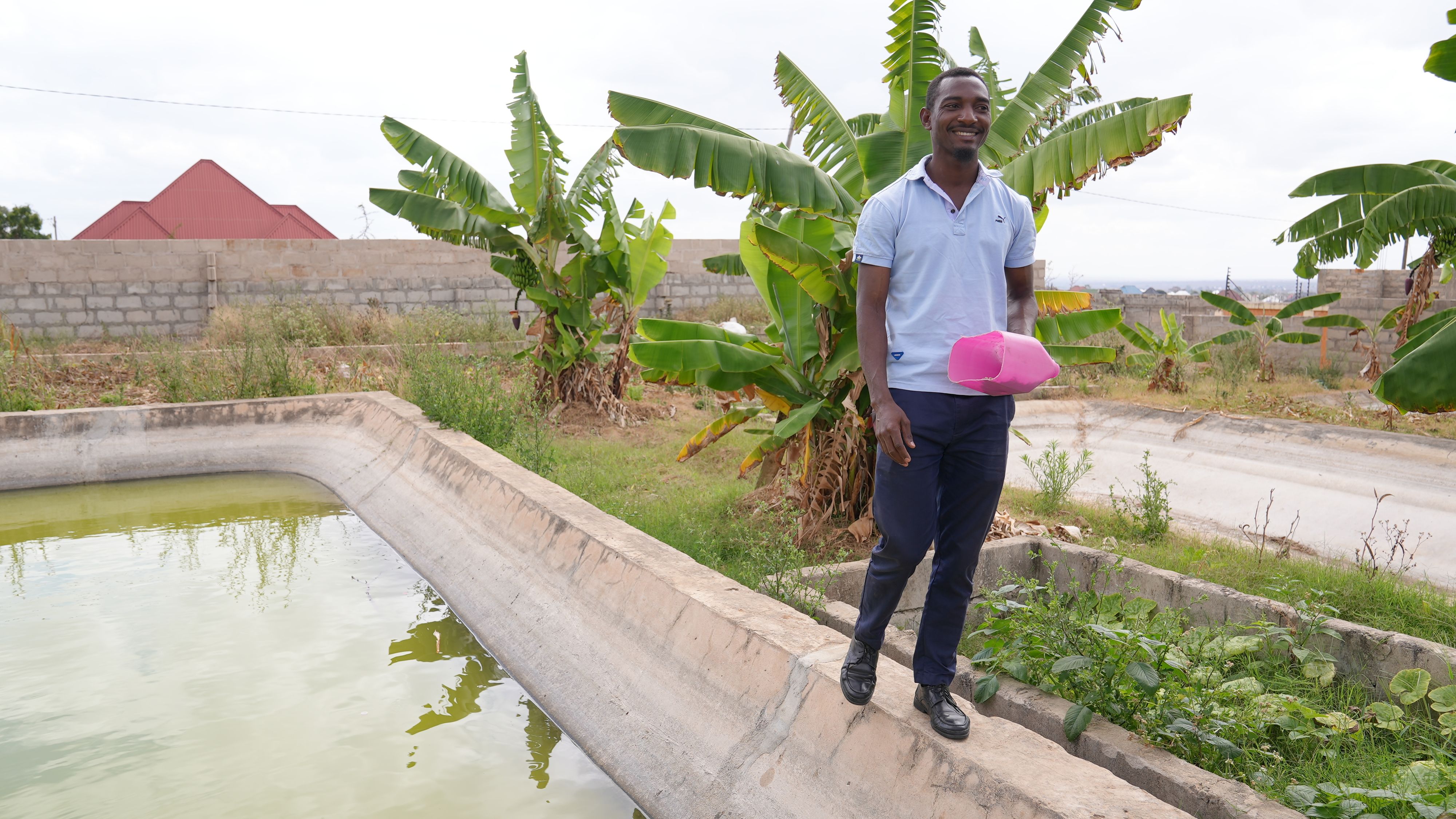Un homme vêtu d’une chemise bleu clair et d’un pantalon bleu marche le long d’un bassin de pisciculture. Il tient un récipient rose dans sa main gauche.