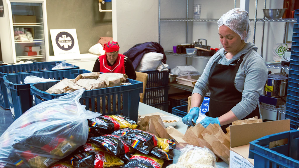 Deux femmes emballent des aliments. Des paquets de pâtes sont posés sur une table au premier plan.