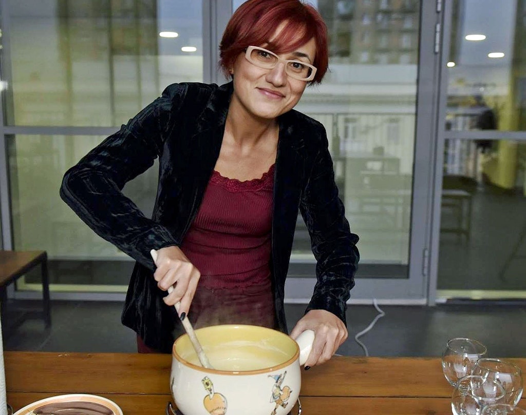 Une femme aux cheveux roux et portant des lunettes remue le fromage dans un caquelon à fondue. 