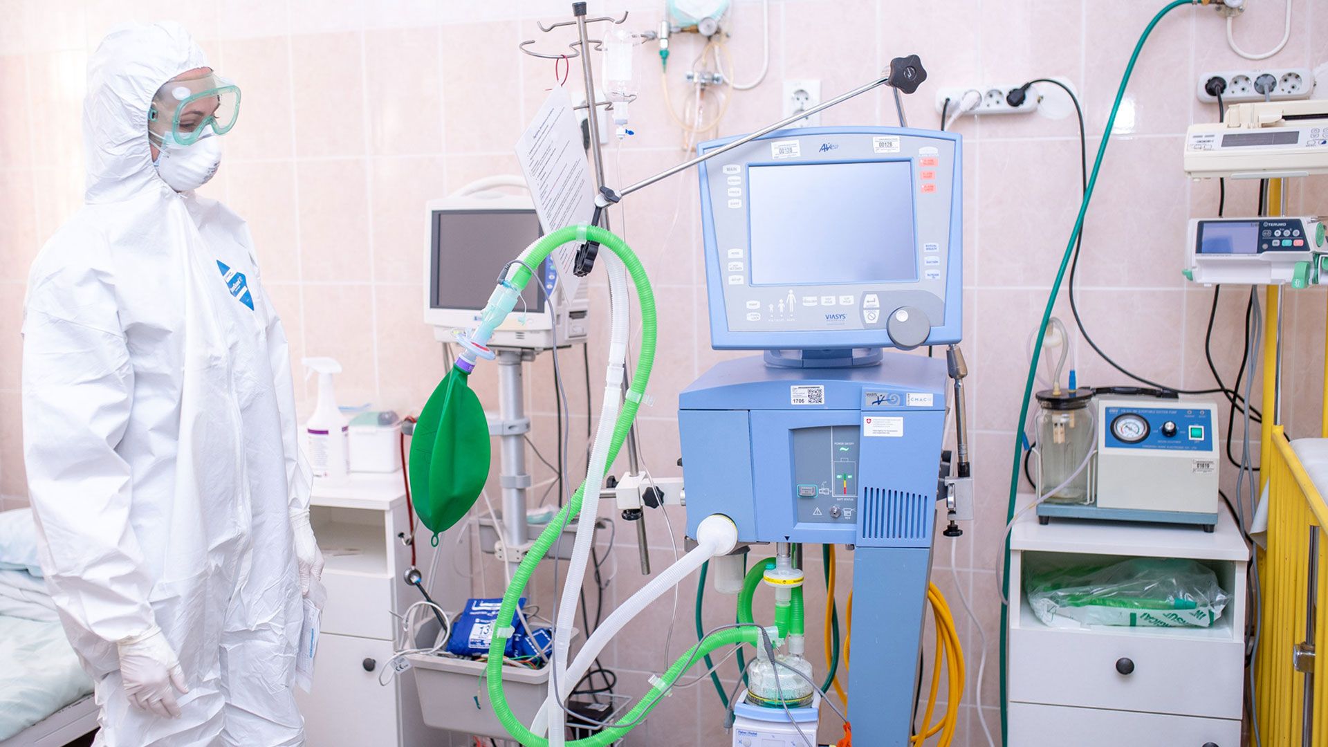Dans une chambre d’hôpital, une femme portant un masque, des lunettes et une combinaison de protection se tient debout à côté d’appareils médicaux.