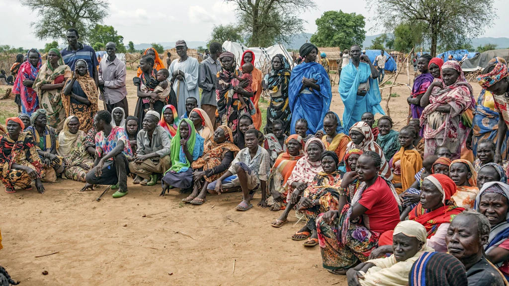Plusieurs dizaines de personnes, hommes, femmes et enfants se tiennent debout ou sont assises directement sur le sol en terre