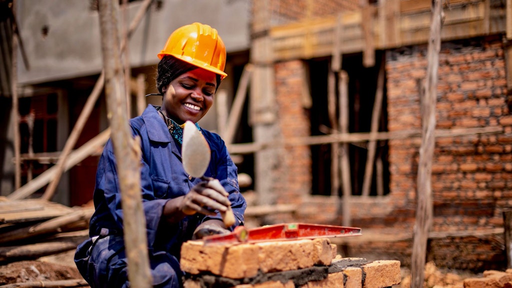 Une femme maçonne des briques sur un chantier de construction, devant un bâtiment en cours d’édification.