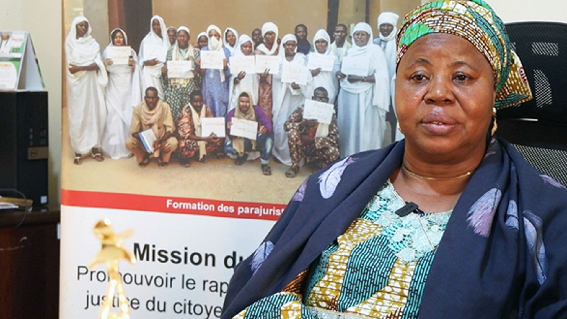 La directrice exécutive de WILDAF Mali est assise sur une chaise de bureau. Sur le fond, il y a une affiche avec des femmes artisans de paix