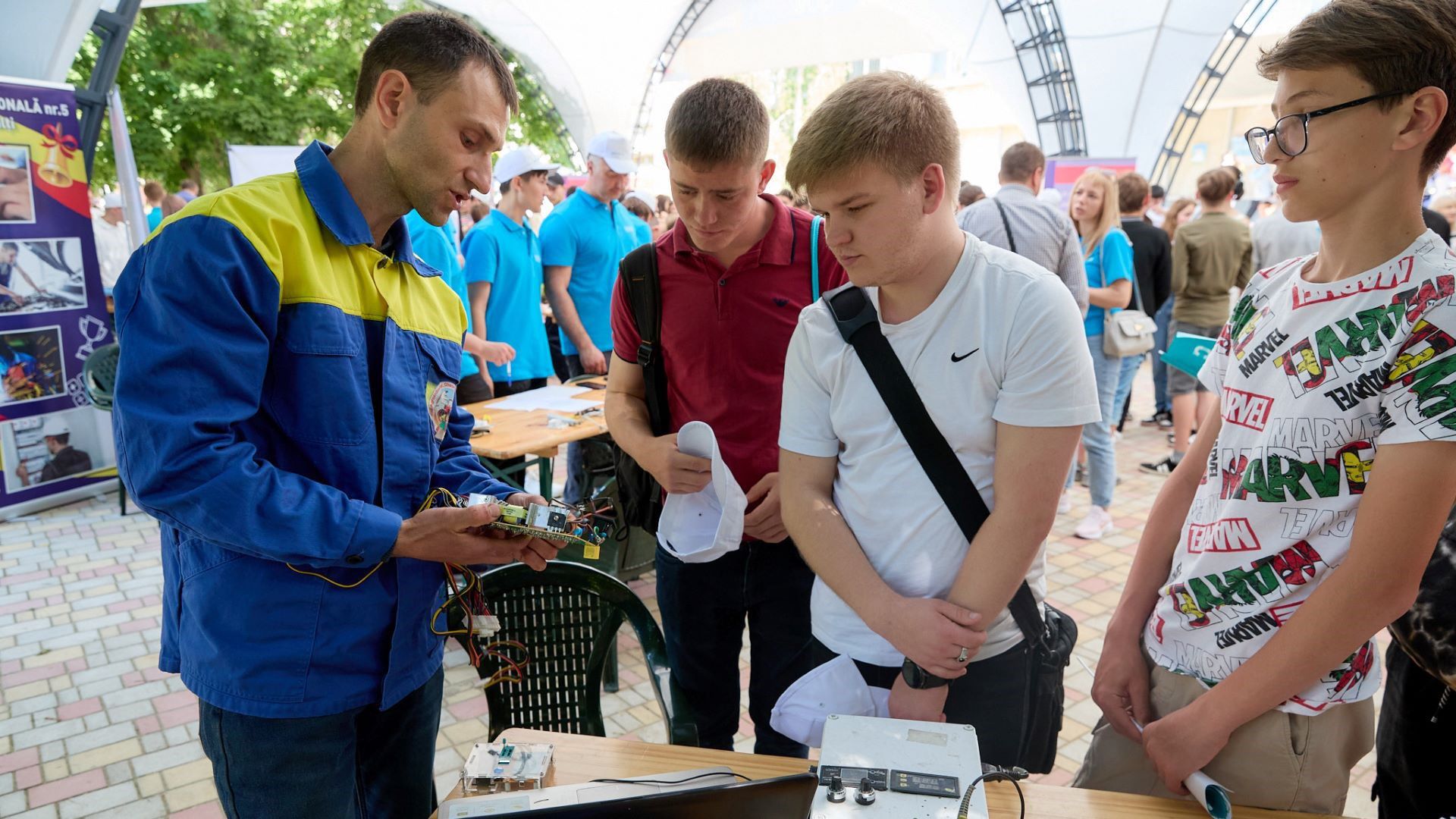 Un homme vêtu d’une veste bleu et jaune montre un appareil électronique à trois adolescents lors d’un salon professionnel.