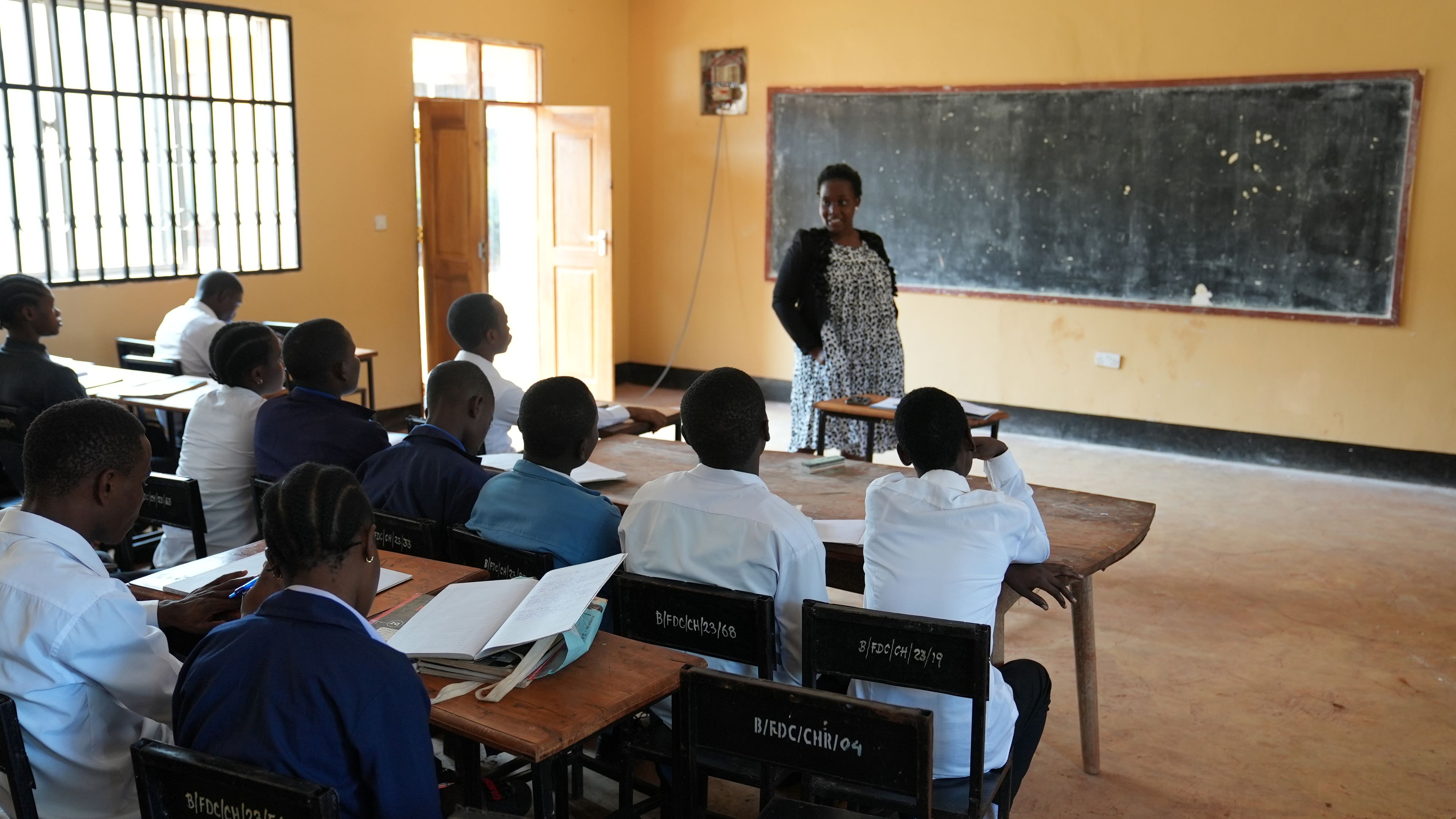 Des élèves sont assis à leur bureau, de dos. Une enseignante se tient devant la classe, les mains dans les poches.