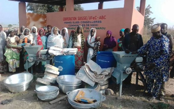 Un groupe de femmes portant des bébés et trois hommes se tiennent devant un bâtiment où il est écrit «Coopération suisse – Femmes élues conseillères communales de Nikki».