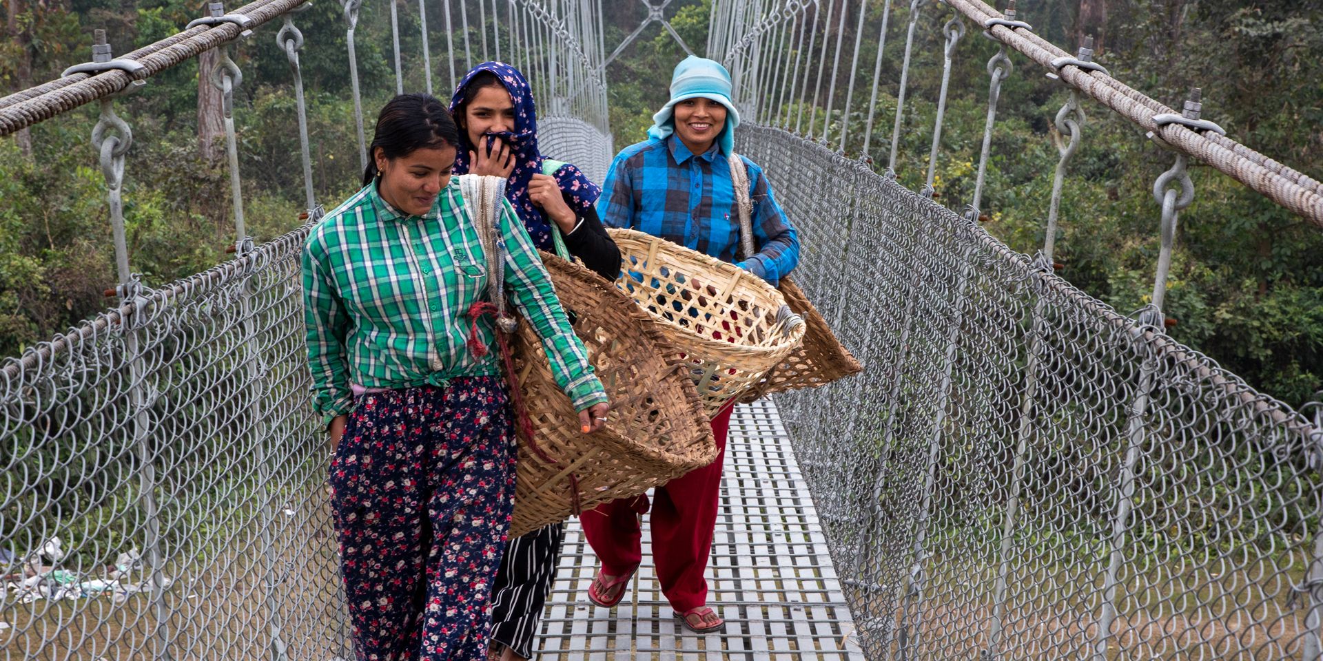 Trois femmes népalaises traversent un pont suspendu. Elles portent de grands paniers tressés vides.