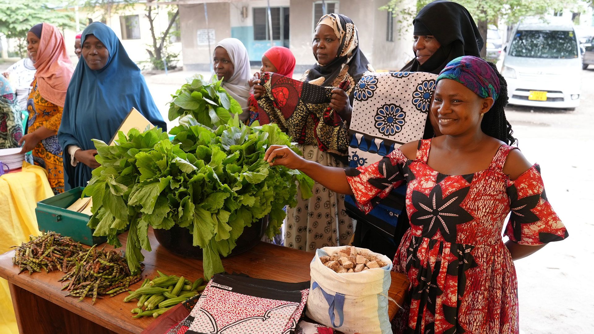 Sei donne in piedi intorno a un tavolo. Sul tavolo vi sono una grande quantità di lattuga, un sacco bianco pieno di pietre, indumenti colorati, del gombo e dei piselli. 