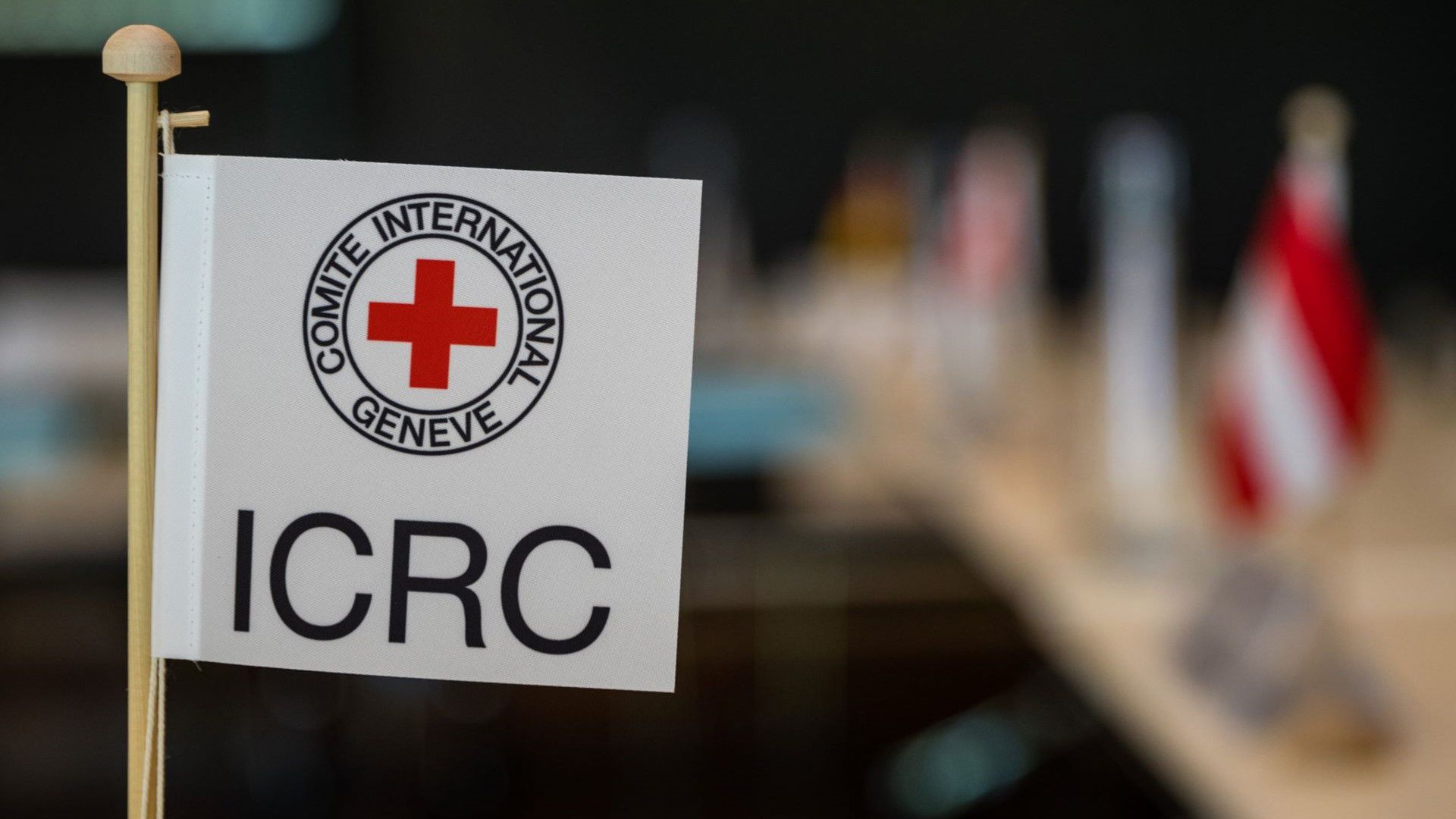 Bandiera da tavolo con il logo del Comitato Internazionale della Croce Rossa. Sullo sfondo si riconoscono vagamente altre bandiere e un tavolo da conferenza.