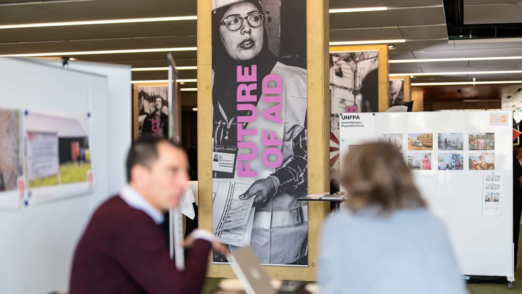 Due persone parlano sedute a un tavolo. Sullo sfondo, un pannello mostra il tema dell’IC Forum: il futuro dell’aiuto umanitario.