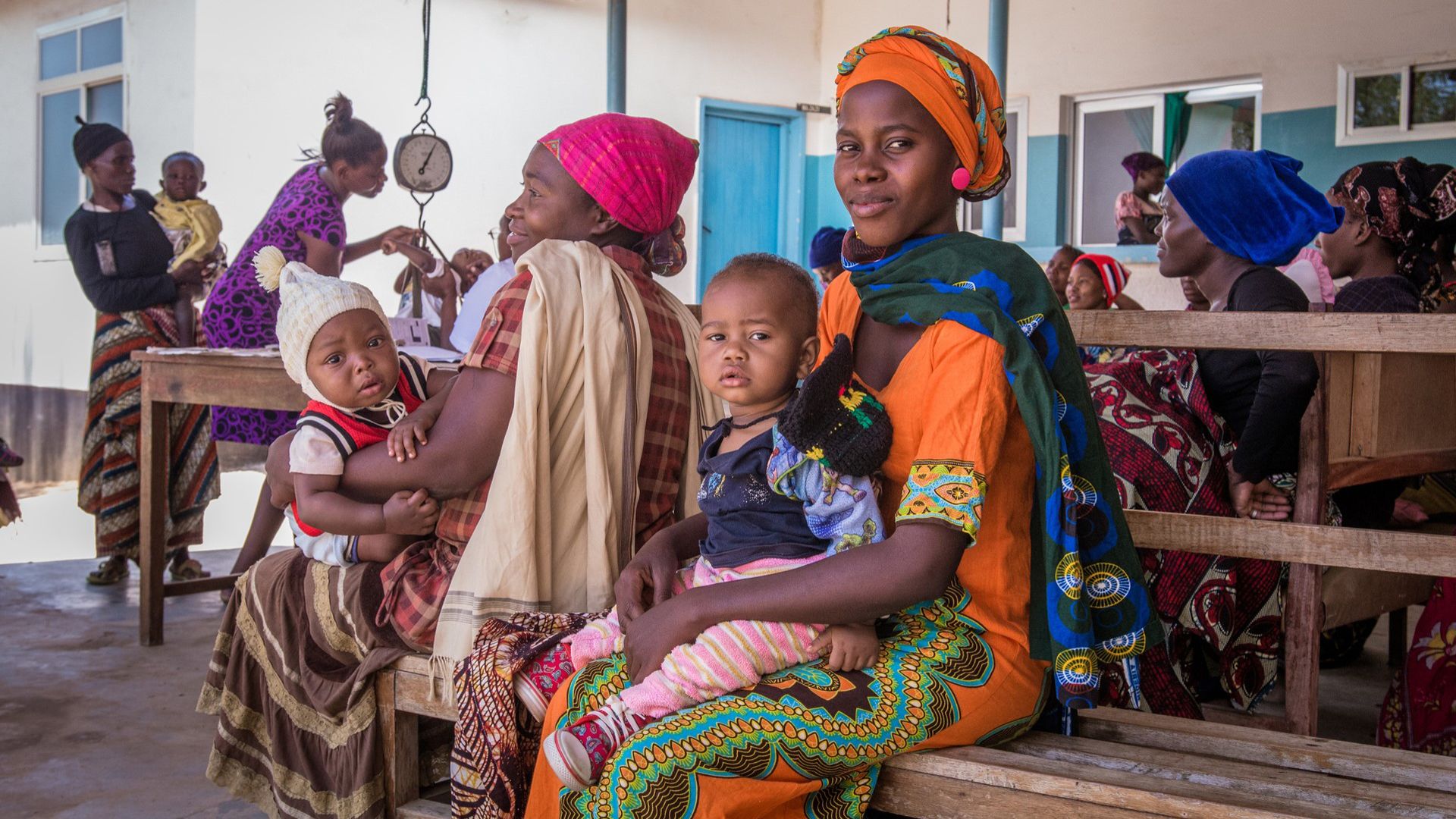 Donne sedute su una panca con i bambini in grembo presso un centro sanitario tanzaniano; sullo sfondo una bilancia.