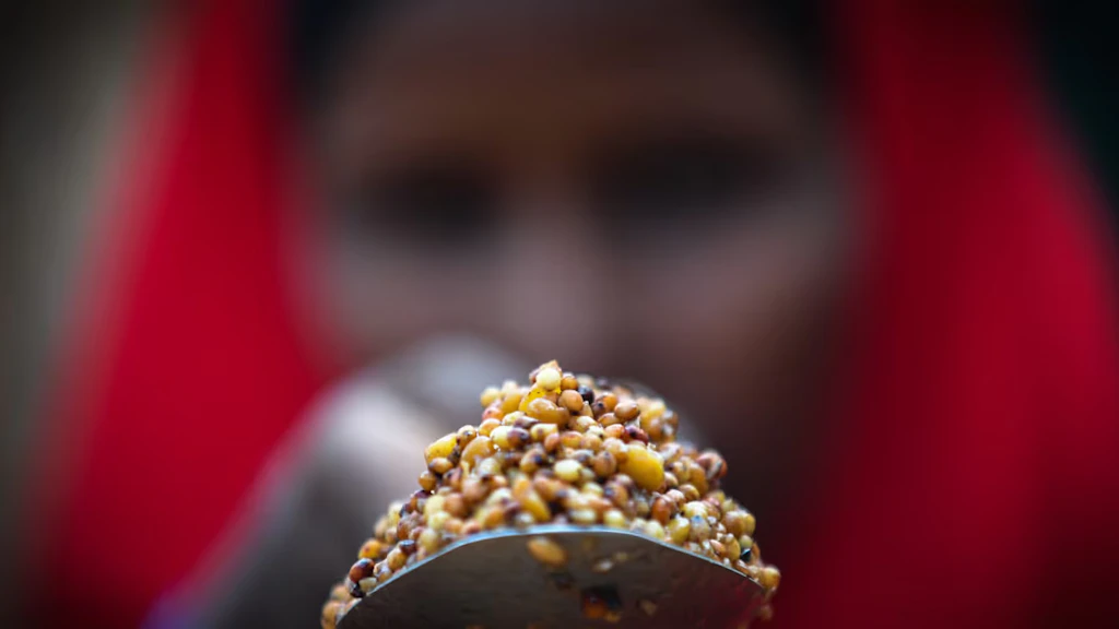 Ritratto di donna in primo piano con sciarpa rossa sullo sfondo. In primo piano, la donna punta con la mano destra un cucchiaio di cereali gialli, marroni e neri verso l'obiettivo della macchina fotografica.