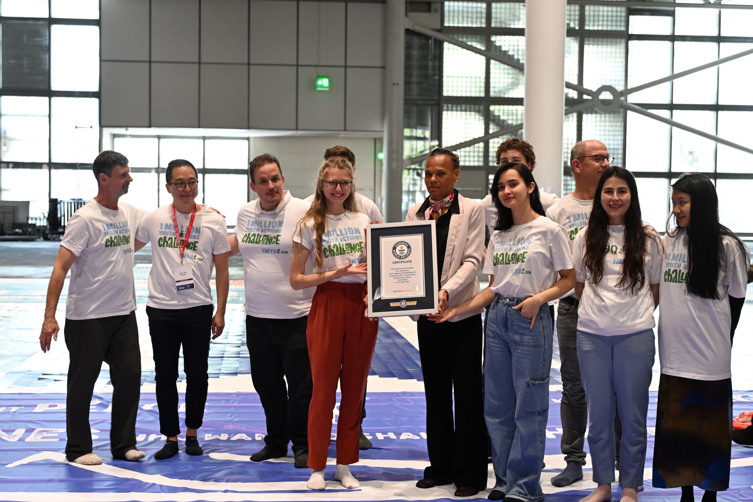 11 persone posano per una foto, indossando T-shirt con la scritta '1 Million Youth Action Challenge'. Al centro, Patricia Danzi, la direttrice della DDC, tiene un certificato del Guinness dei Primati.