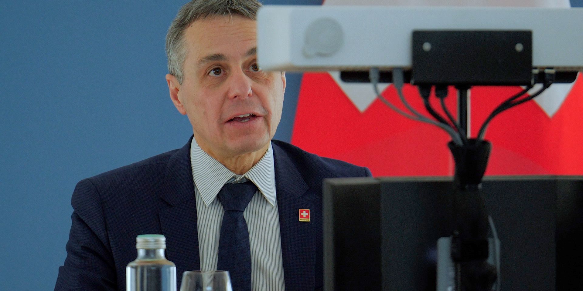 Il consigliere federale Ignazio Cassis davanti a una telecamera durante una conferenza virtuale, la bandiera svizzera in sottofondo.