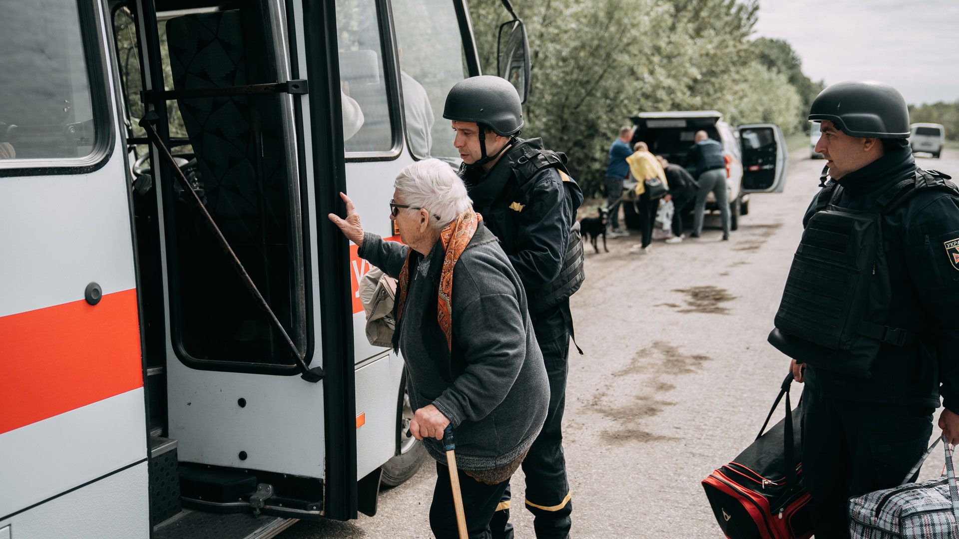 Persone volontarie aiutano una donna anziana con il bastone a salire su un autobus di evacuazione lungo una strada di campagna.